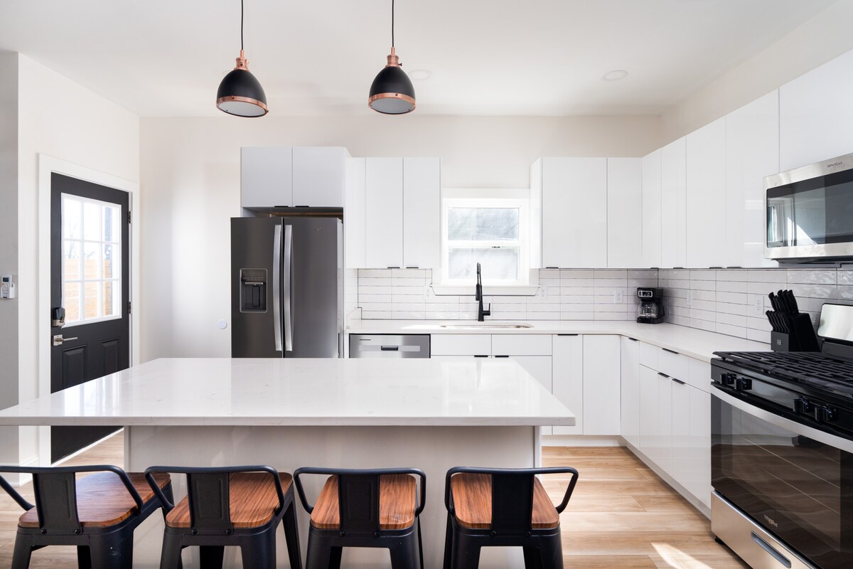 A modern kitchen is equipped with sleek white cabinetry and stainless steel appliances. A large island with a white countertop provides ample workspace. Six black metal chairs are arranged around the island. Natural light enters through a nearby window, enhancing the bright atmosphere.