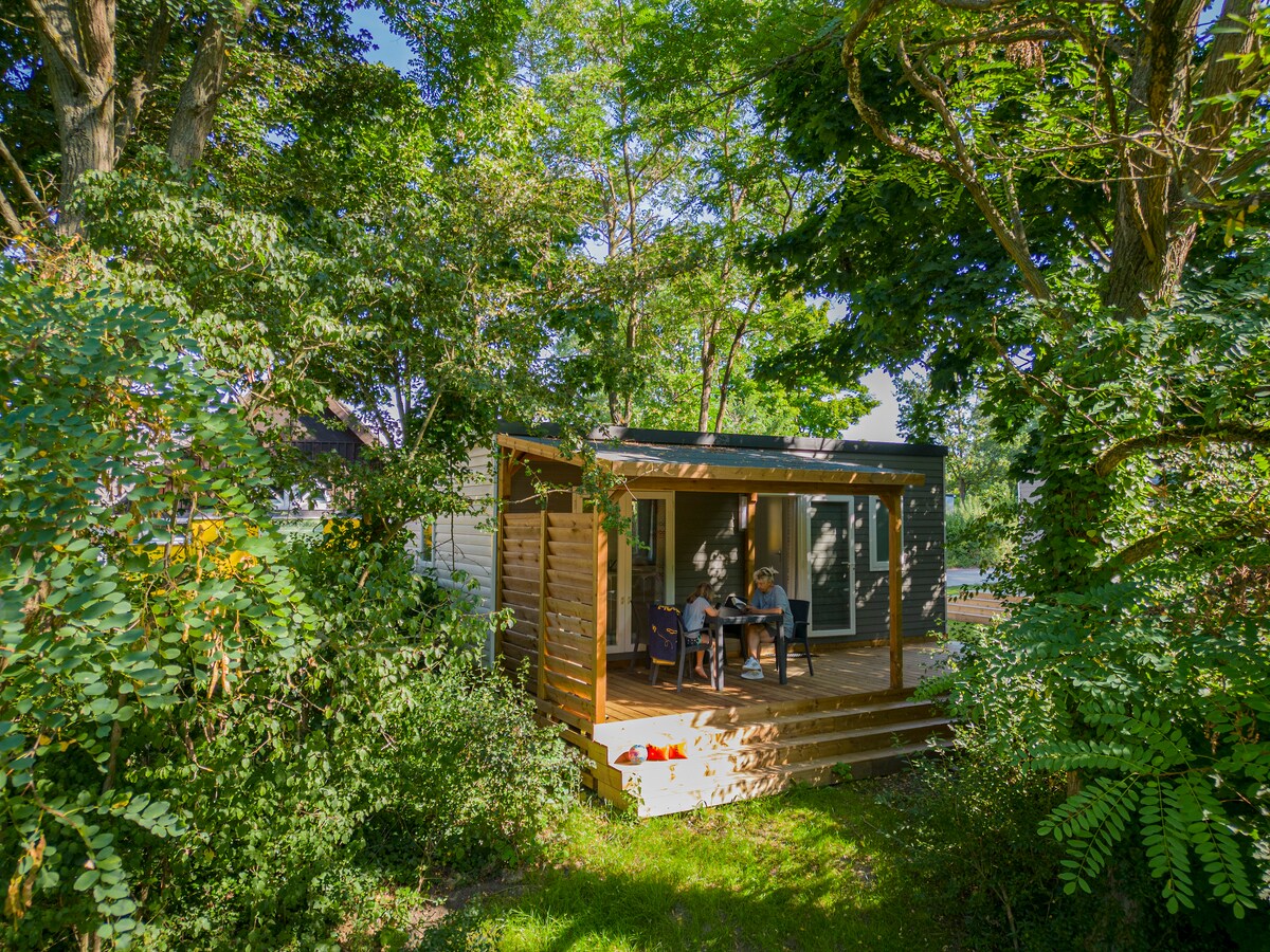 A cozy mobile home is nestled among the lush greenery, providing a sense of privacy. A wooden deck is visible, featuring seating for two, surrounded by tall trees and vibrant foliage, creating a serene outdoor retreat.