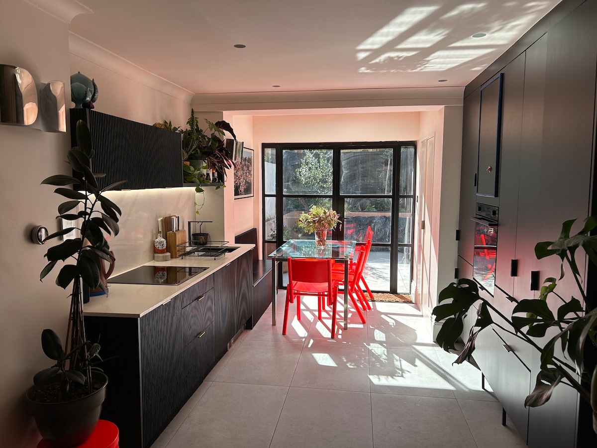A modern kitchen area features sleek black cabinetry with a white countertop and integrated appliances. A glass dining table is surrounded by red chairs, creating a vibrant contrast. Large windows allow natural light to fill the space, highlighting indoor plants placed throughout.