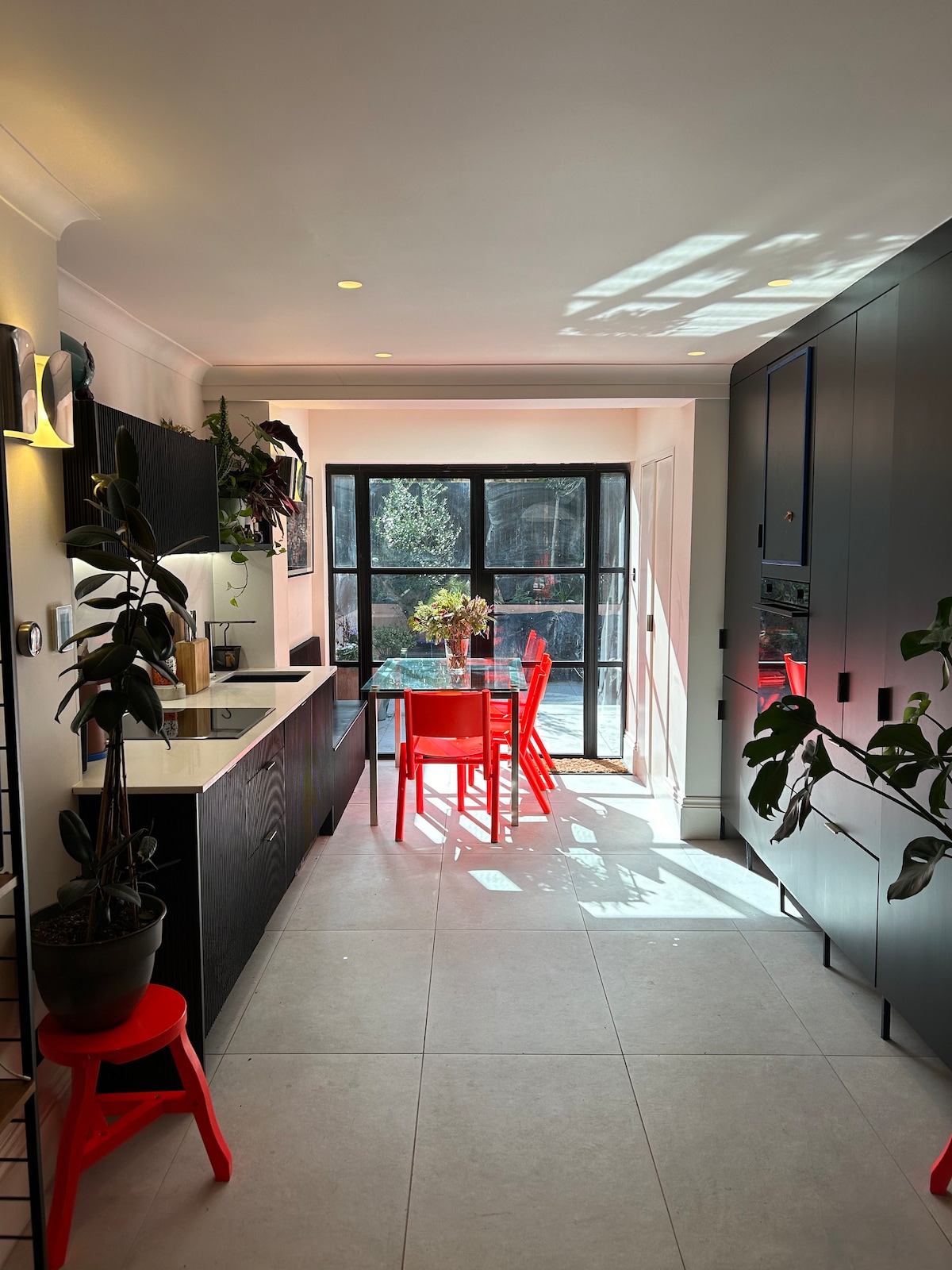 A bright kitchen showcases sleek black cabinetry and a modern countertop. A glass dining table surrounded by vibrant red chairs is positioned near a large window, allowing natural light to filter through. Potted plants add a touch of greenery to the space.