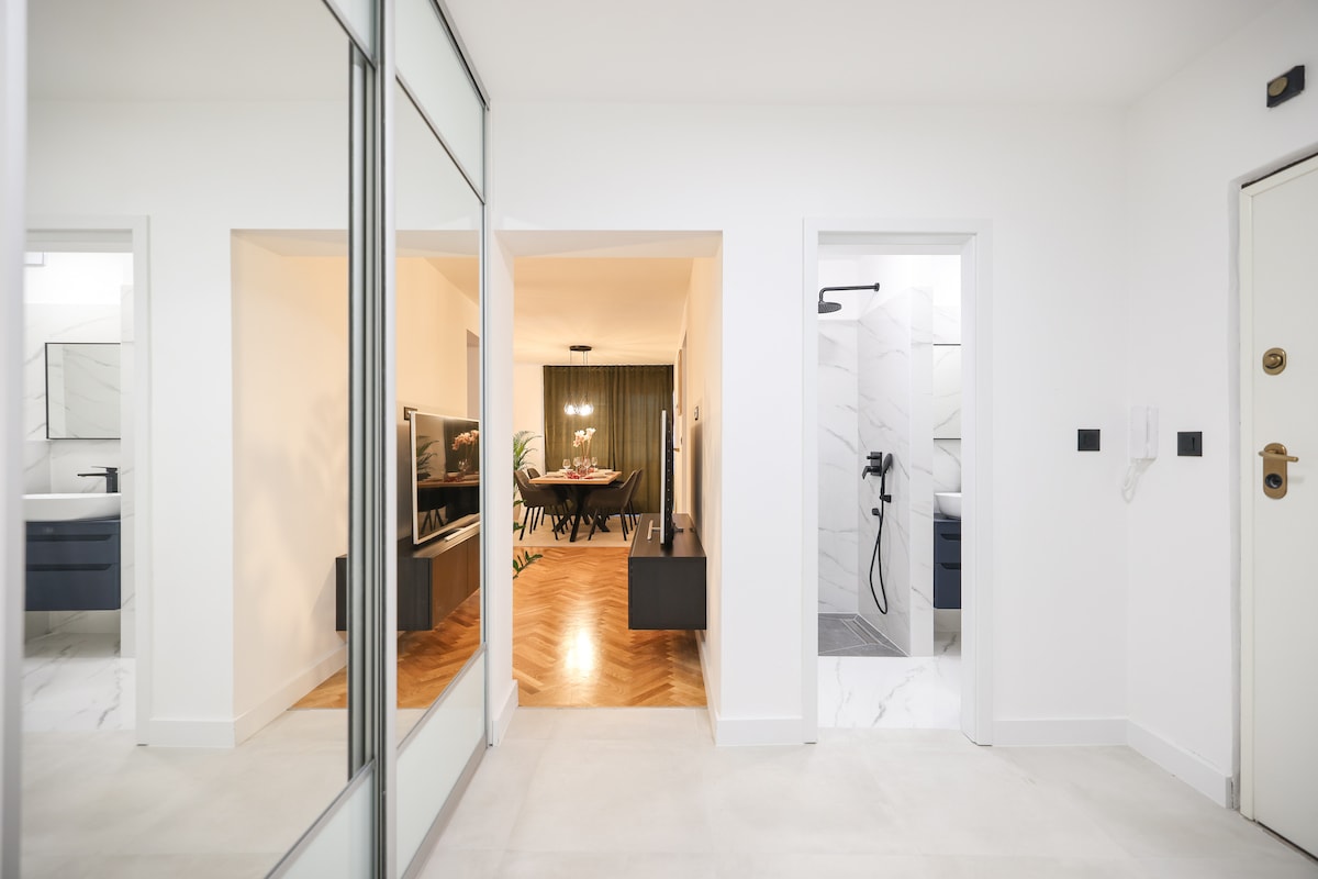 A bright hallway with glossy white walls is seen, featuring two doorways leading to different areas. One doorway opens to a modern bathroom with a walk-in shower, while another leads to a spacious living and dining area, with large windows allowing natural light.