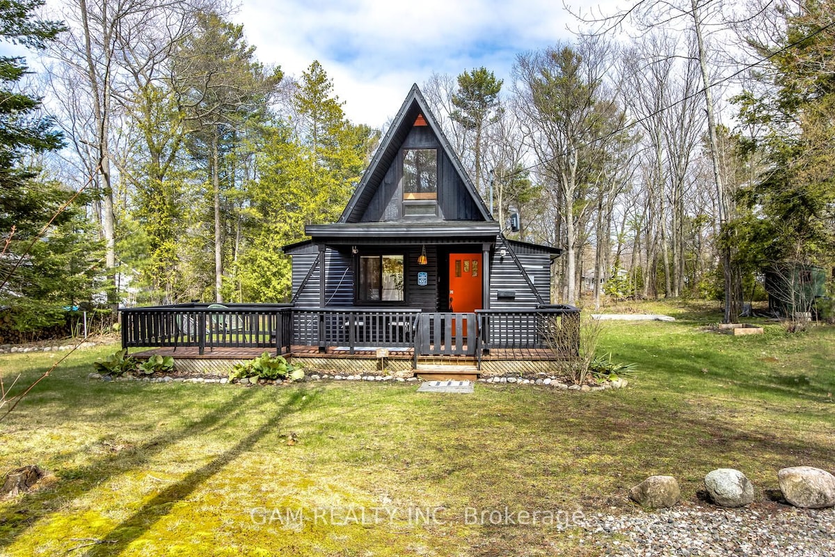 A newly updated A-Frame cottage is set amidst lush greenery, featuring a dark exterior and a prominent orange front door. A wooden deck wraps around the front, surrounded by a landscaped yard with decorative stones and a variety of trees in the background.