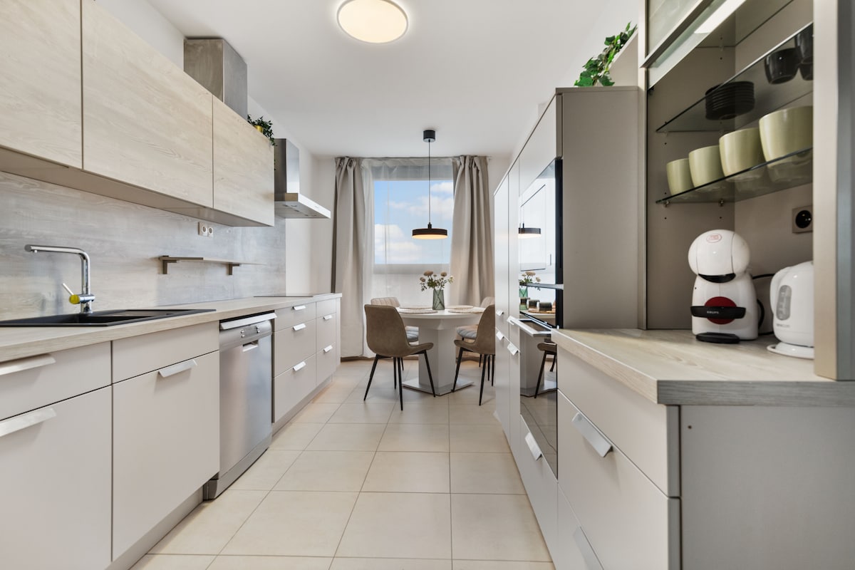 A modern kitchen is presented with sleek cabinetry and stainless steel appliances. A small dining area is featured with two chairs and a circular table, positioned near a window adorned with light curtains. Soft natural light enhances the inviting ambiance.