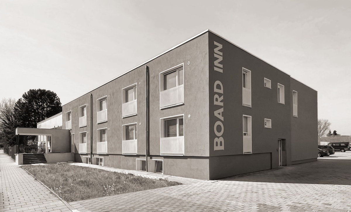 The exterior of a modern boarding house is depicted, featuring a minimalist design with a large sign displaying 'BOARD INN'. The building includes multiple windows arranged symmetrically, and a clean pathway leads to the entrance, surrounded by green grass.