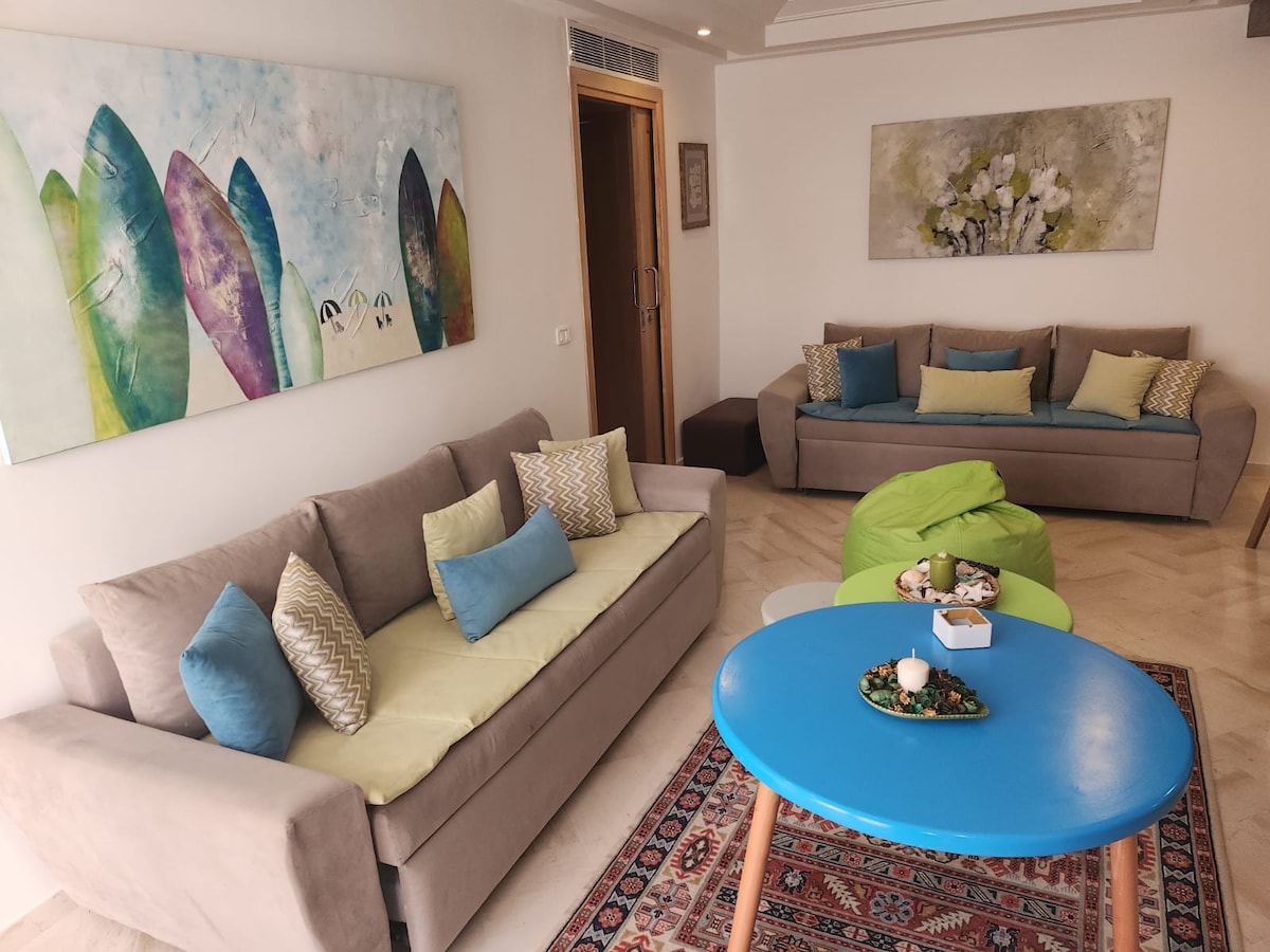 A lively living area features soft, neutral sofas adorned with blue and green cushions. A round, blue coffee table is centered between the seating, which is accompanied by a patterned rug. Colorful wall art depicting surfboards and floral designs enhances the space.