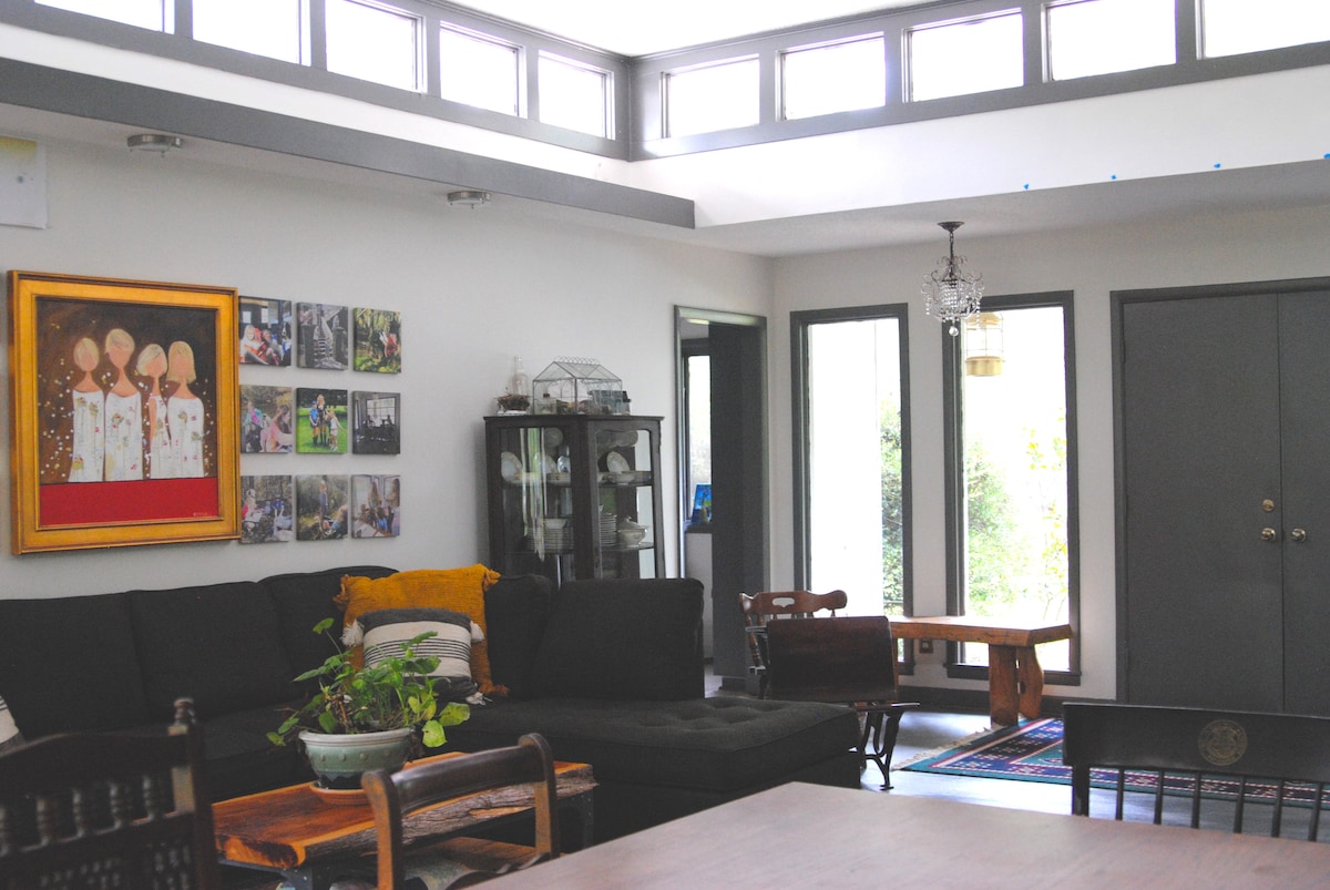 A spacious living area features large windows allowing natural light to fill the room. Comfortable seating is arranged around a low coffee table, while a mix of artwork adorns the walls. A glass display cabinet stands in one corner, showcasing decorative items.