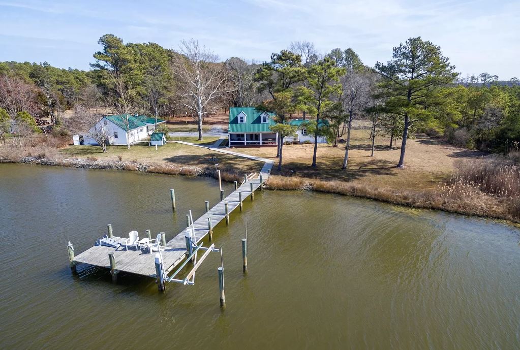 A private dock extends into the calm waters of St. John's Creek, surrounded by trees and grassy areas. Two distinct houses are visible nearby, featuring green roofs and ample outdoor space, creating a serene setting ideal for relaxation and outdoor activities.
