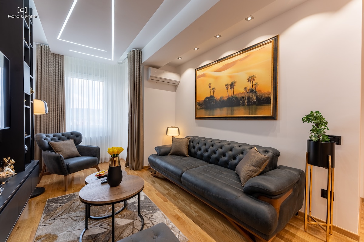 A modern living room is presented with a sleek leather sofa and two matching armchairs. A round coffee table sits in the center, adorned with a decorative vase and a small plant. Soft light is provided by lamps beside the seating area, while a large artwork decorates the wall.