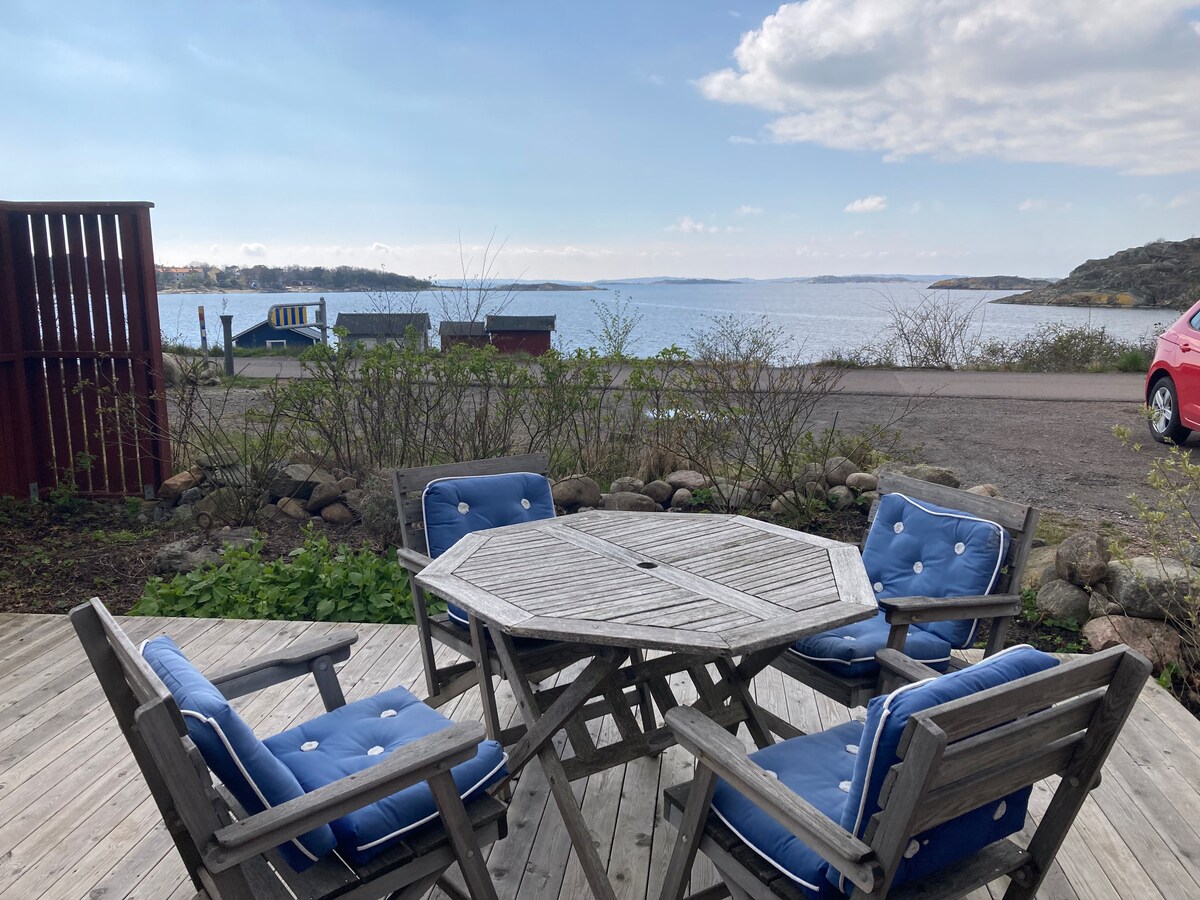An outdoor seating area is featured with a wooden octagonal table surrounded by six blue cushioned chairs. The view includes a calm body of water and distant greenery, with a car visible nearby and a red wooden fence framing the scene.