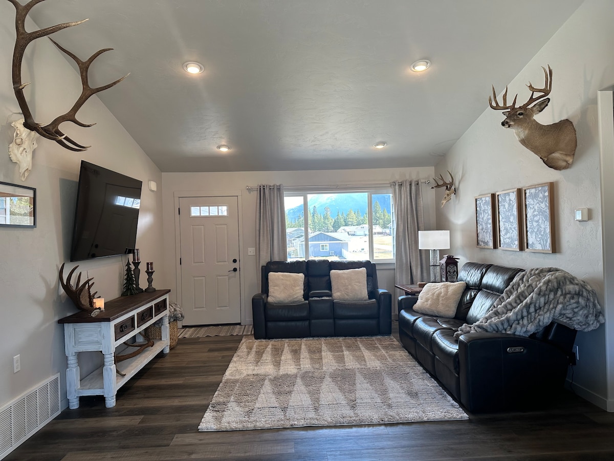 A cozy living room features two leather sofas with soft cushions, offering a relaxed seating area. A television is mounted on the wall, while natural light filters in through the windows. Antler decor adds a rustic touch, and a textured area rug defines the space.