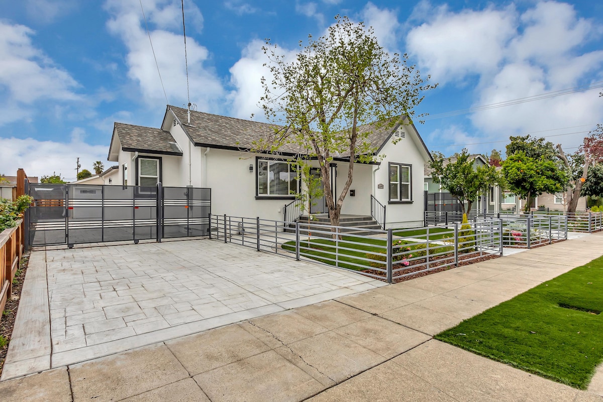 A modern house is presented with a well-maintained exterior, featuring a spacious patio area bordered by a sleek metal fence. Lush greenery and trees accentuate the front yard, while large windows allow natural light to fill the interior.
