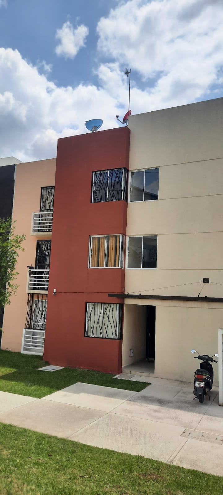 A two-story building with a combination of light and dark exterior colors is depicted. The structure features several windows with decorative metal grilles. A grassy area is visible in the foreground, providing a welcoming entrance to the residence.