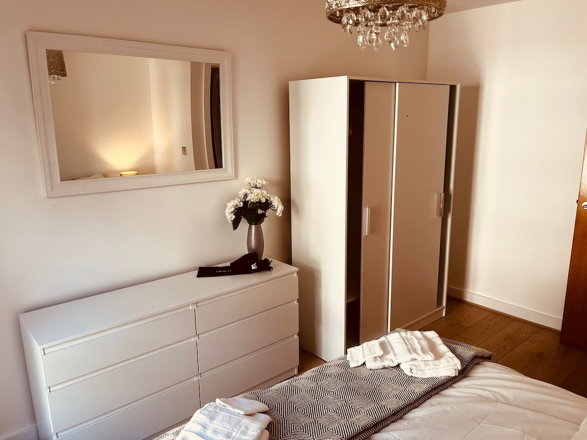 The bedroom features a dresser and a wardrobe with a clean white finish. A mirror reflects soft lighting, enhancing the airy atmosphere. A neatly made twin size bed is positioned beside the furniture, with a cozy blanket and towels placed on top.