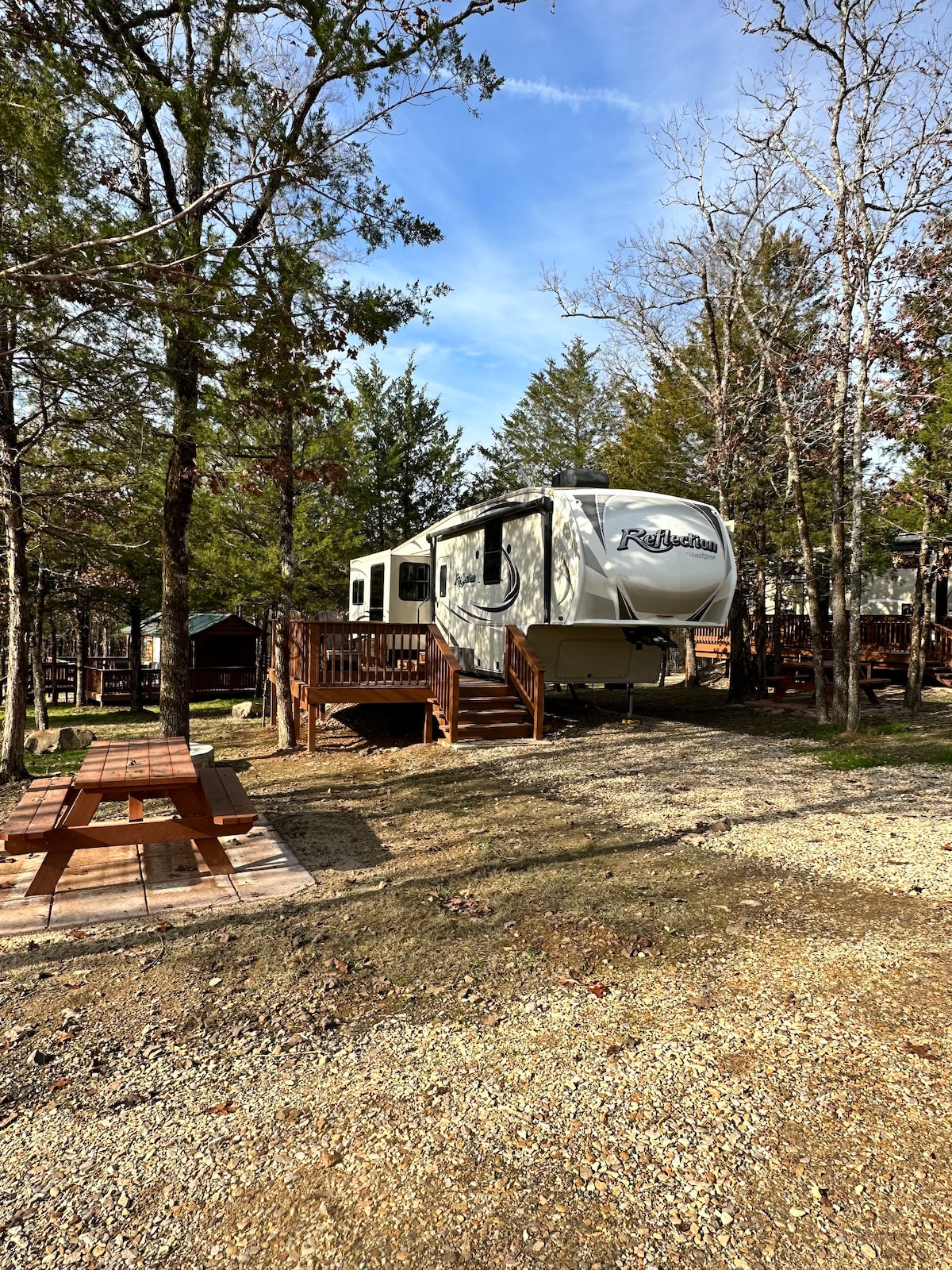 A spacious trailer sits on a gravel area surrounded by trees. A wooden deck leads to the entrance, while a picnic table is positioned on a wooden platform nearby, inviting outdoor dining. Natural light filters through the foliage, creating a serene atmosphere.