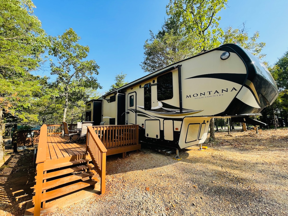 A spacious RV is parked on a gravel surface, surrounded by trees. A wooden deck with a railing leads to the entrance, offering easy access. The exterior features a cream and black color scheme, complemented by large windows for natural light.