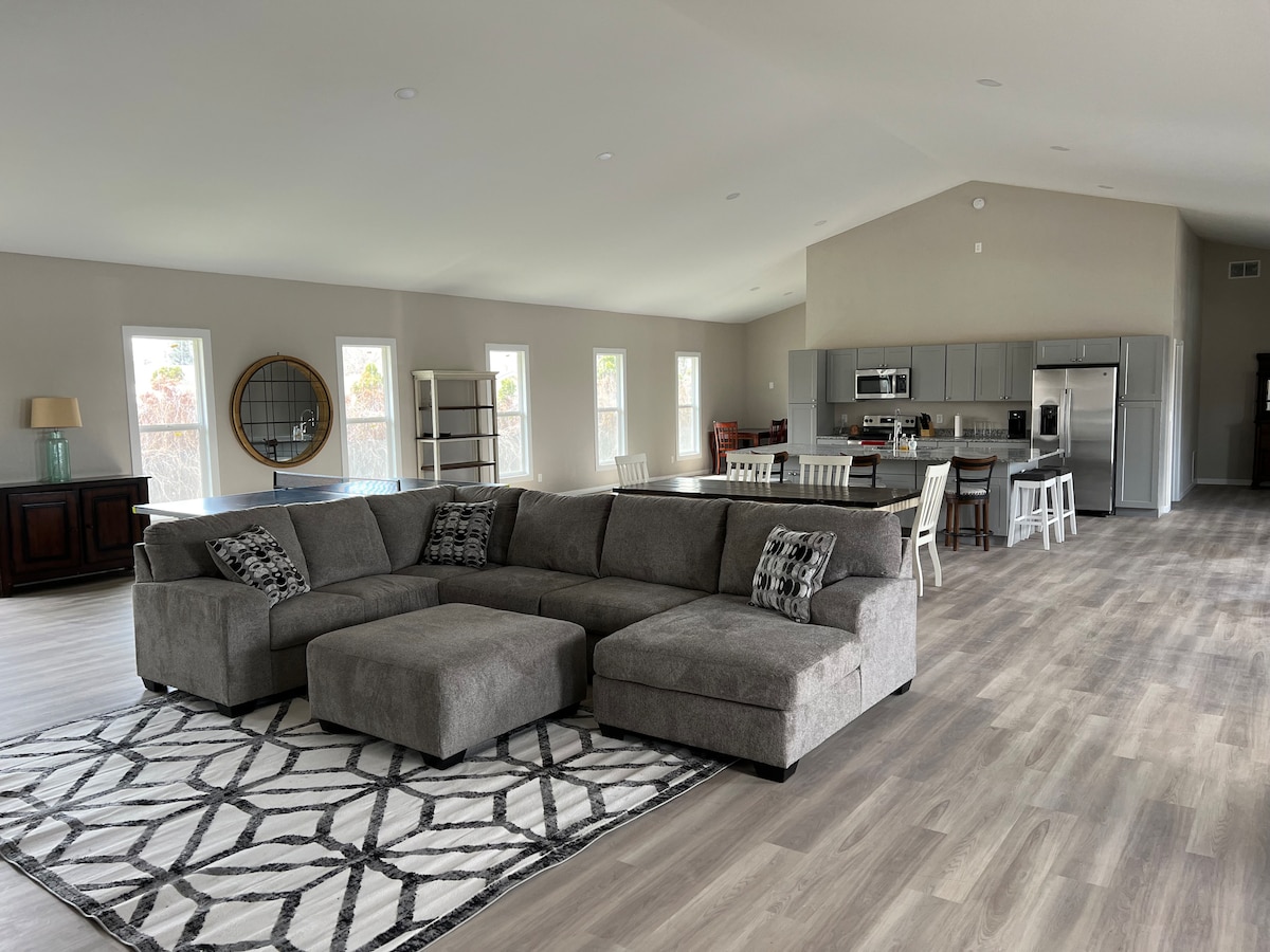 An expansive living area is highlighted by a large sectional sofa arranged on a patterned rug. Sunlight enters through several windows, illuminating the neutral-toned walls. A dining area with a table and chairs is positioned on one side, while a modern kitchen is visible in the background.