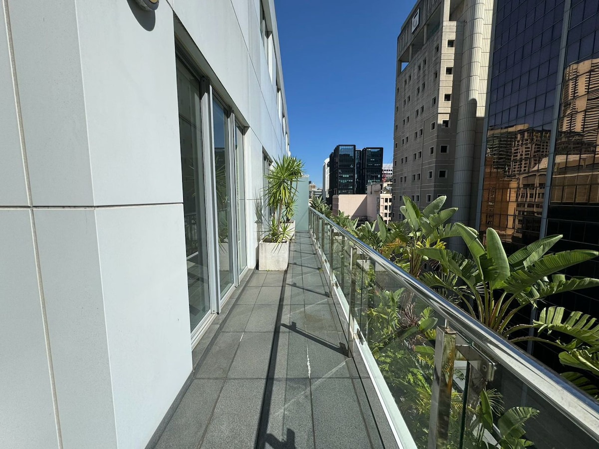 A spacious balcony extends alongside the apartment, featuring glass panels that offer unobstructed views of the city skyline. Lush green plants in planters add a natural touch, enhancing the outdoor space. The clear blue sky provides a bright backdrop to this inviting area.