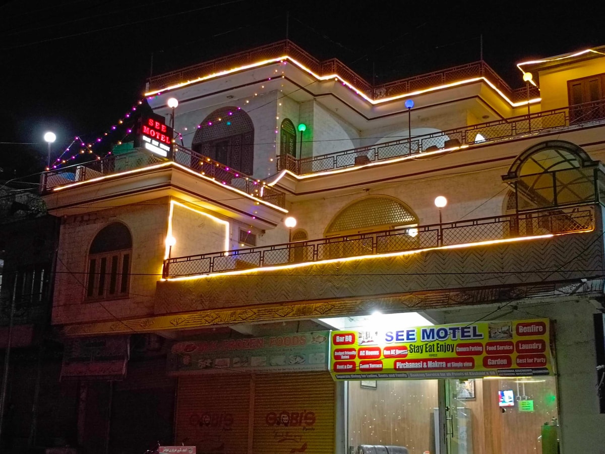 The exterior of a well-lit motel is showcased at night, with colorful lights outlining the building's architecture. A sign reading 'SEE MOTEL' is prominently displayed, while illuminated decorative elements add vibrancy. A storefront featuring a variety of services is visible at the base.