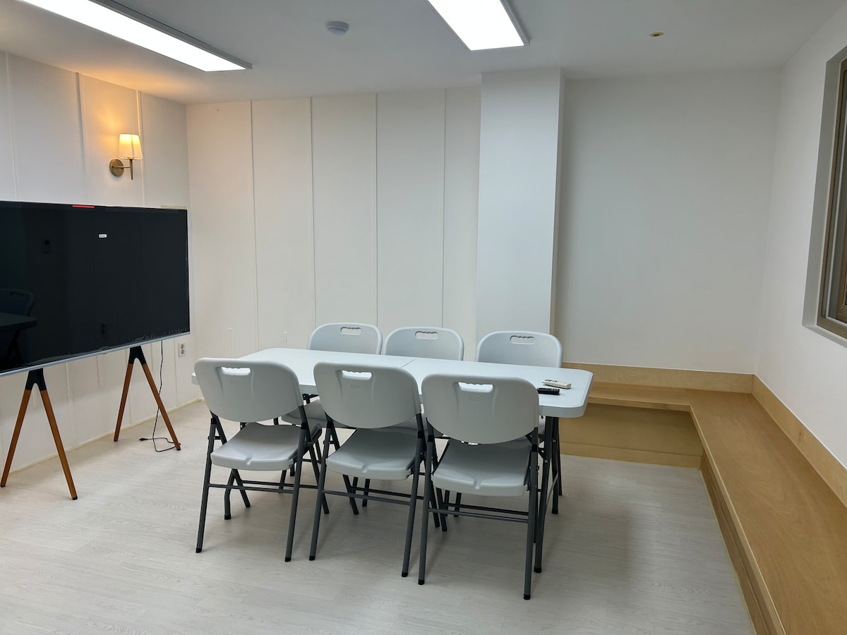 A bright meeting space is presented, featuring a rectangular table surrounded by six folding chairs. A large whiteboard on an easel is positioned to one side, while a wooden bench runs along the wall, enhancing the room's functionality.