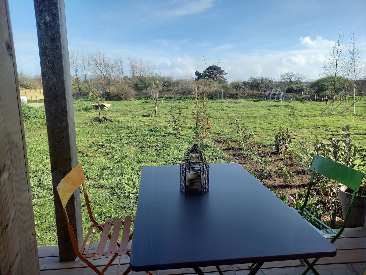 A garden view is captured from a covered terrace, featuring a dark table with a decorative lantern at its center. Surrounding the table are brightly colored chairs in orange and green. The expansive green lawn is dotted with young trees and shrubs under a clear blue sky.