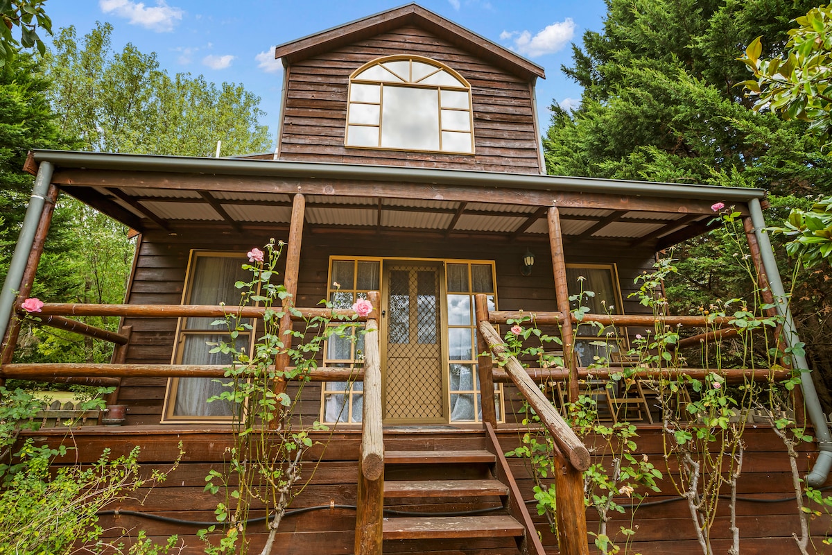 The quaint cottage features a rustic wooden exterior complemented by a wraparound porch. A set of steps leads to double doors, framed by tall windows that enhance natural light. Lush greenery and rose bushes accentuate the surroundings, providing a serene entryway to the home.