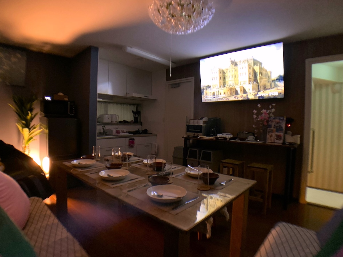 A dining area is situated in an inviting space, with a wooden table set for a meal. Four plates and glasses are arranged neatly. A large screen displays an image, while a modern chandelier provides ambient lighting. The kitchen area is partially visible in the background.