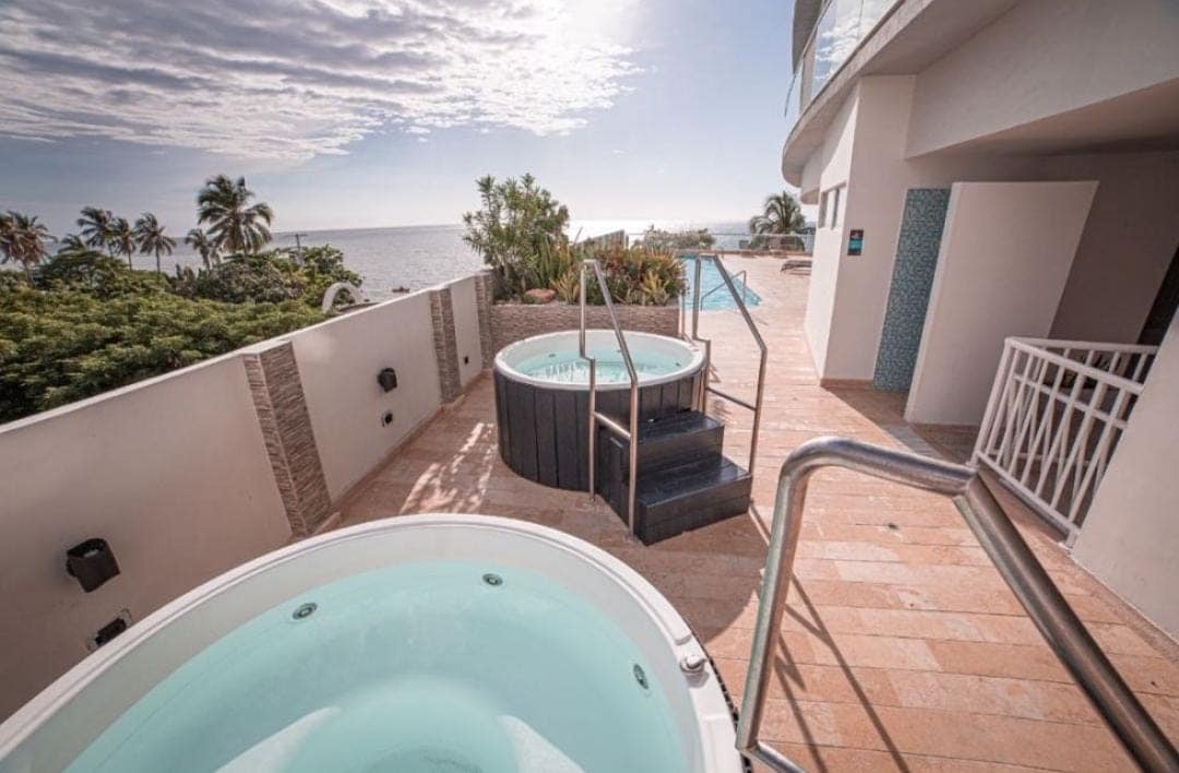 Two outdoor jacuzzis are positioned on a sunlit terrace with a view of the sea. Lush greenery surrounds the area, and a staircase leads to additional amenities. The calm waters of the jacuzzis reflect the nearby palm trees and sky.