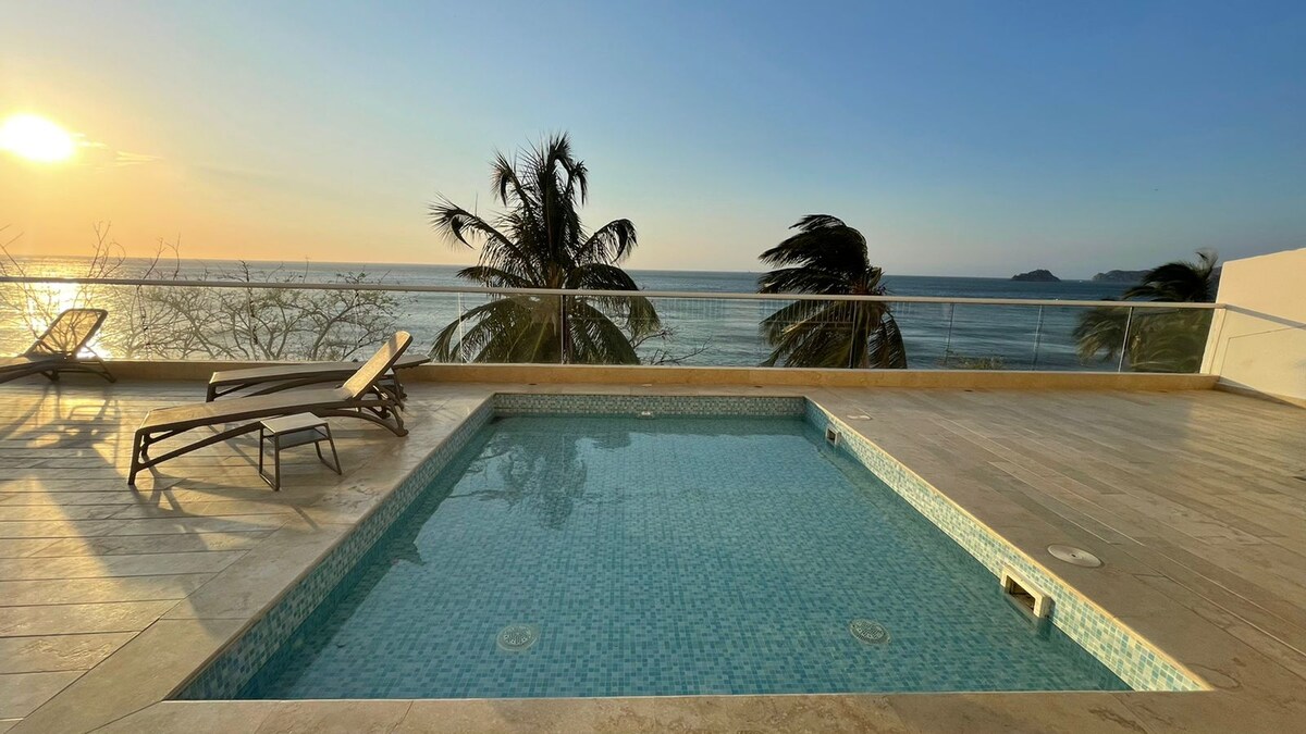 An outdoor terrace features a private pool with clear water, surrounded by a tiled deck. Lounge chairs are positioned nearby, while tall palm trees sway gently in the breeze. The horizon showcases a tranquil sea, illuminated by the soft light of the setting sun.