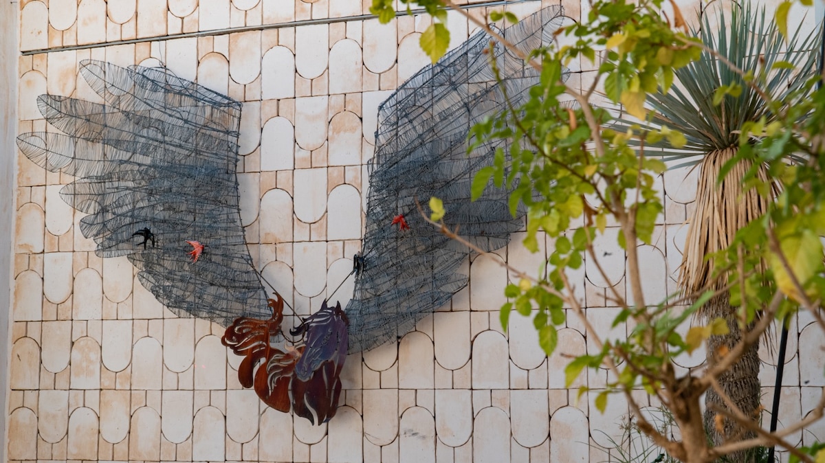 A decorative mural featuring large wire wings extends across a textured wall. The wall is adorned with pale tiles, enhancing the artwork's prominence. Nearby, a green plant adds a touch of nature, contributing to the space's vibrant atmosphere.