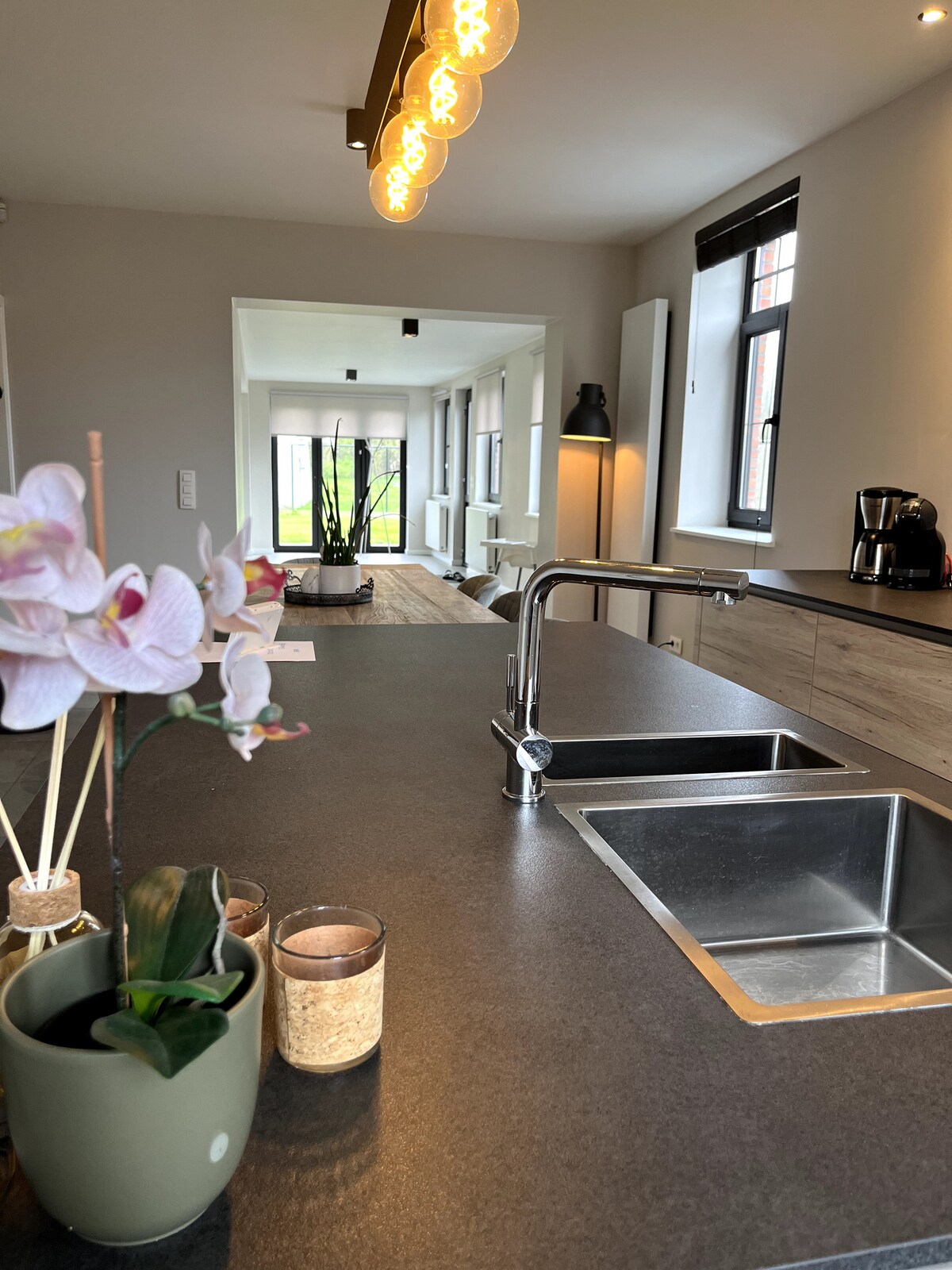 A modern kitchen space features sleek countertops with a double sink and elegant fixtures. Decorative plants and candles are arranged on the counter, while a stylish light fixture hangs above. The open layout leads to a well-lit dining area visible in the background.