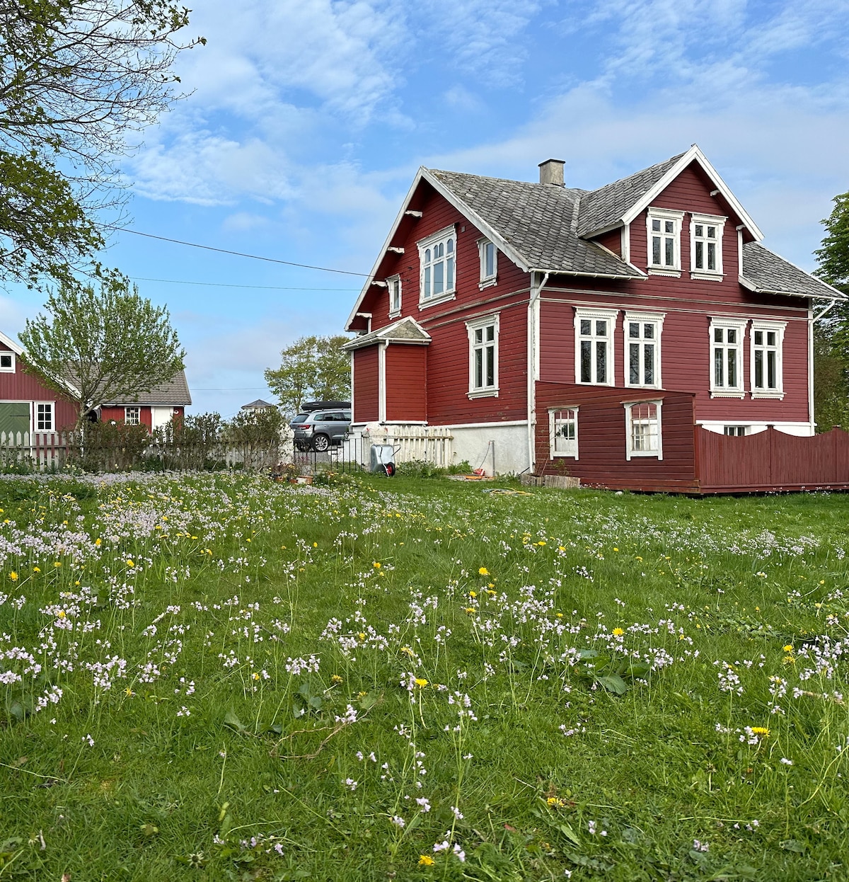 A historic red house with white trim is surrounded by a vibrant green lawn, dotted with colorful wildflowers. The structure features multiple large windows and a steep roof. A serene country atmosphere is enhanced by nearby trees and open sky.