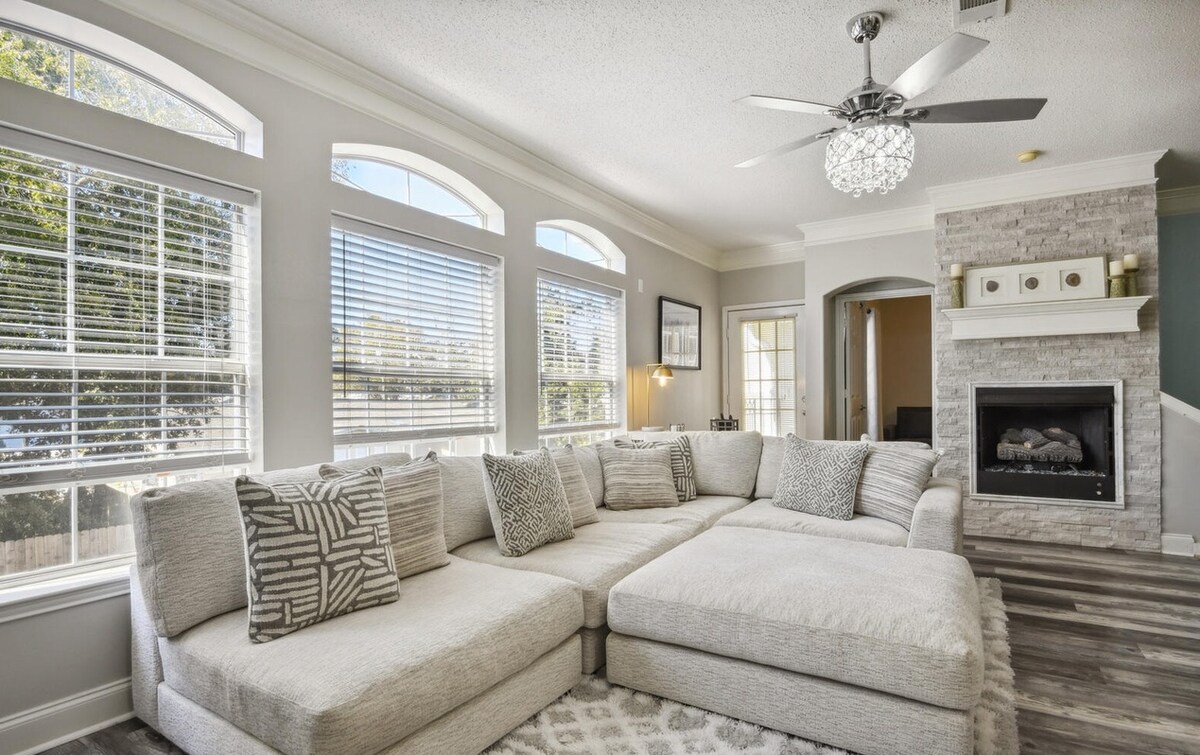 A spacious living area is highlighted by a large sectional sofa adorned with decorative pillows. Natural light flows through multiple arched windows, illuminating the room. A subtle stone fireplace adds a cozy element, while a soft area rug complements the warm tones of the space.