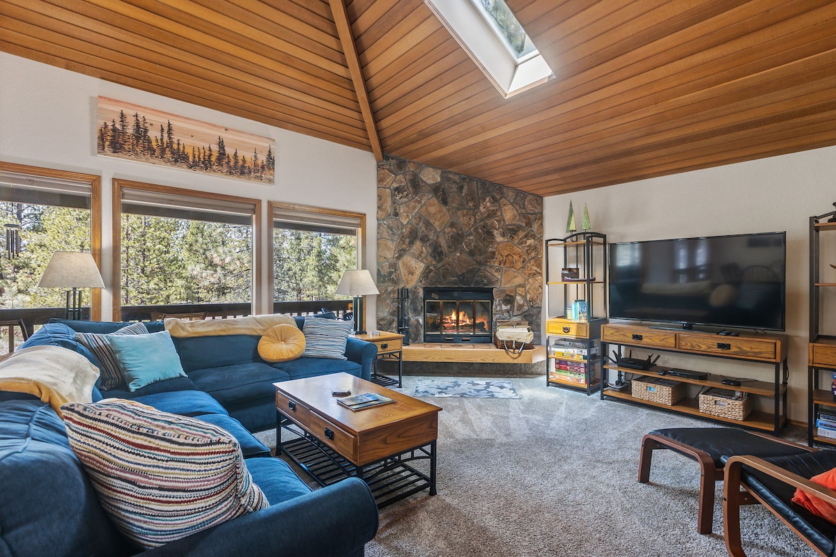 A spacious living room features large windows allowing natural light to flood the area. A cozy sectional sofa is positioned around a wooden coffee table. A stone fireplace is located against a wall with a television mounted nearby. The wooden ceiling adds warmth to the space.
