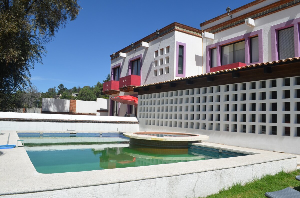 A unique property features an inviting pool area surrounded by landscaped grass. The structure displays a mix of architectural elements, with a vibrant facade and distinctive window designs. The pool is complemented by a circular spa, all under a clear blue sky.