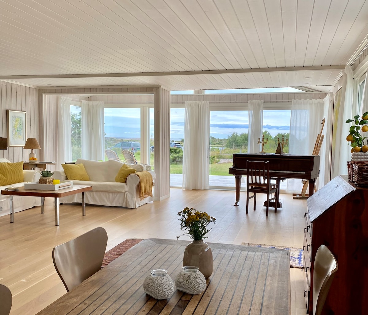 A spacious living area is filled with natural light from large windows, offering views of the serene landscape. A neutral-colored sofa sits alongside a wooden dining table. A piano is positioned thoughtfully in the corner, contributing to a harmonious environment.