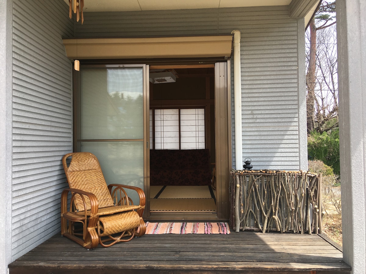 A welcoming entryway features a wooden deck with a woven chair positioned for relaxation. Sliding glass doors open to a traditional interior, with shoji screens casting soft light. A rustic table made of natural materials complements the outdoor space, surrounded by greenery.