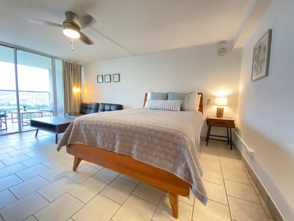 A spacious studio features a comfortable bed with a textured blanket. A sectional sofa in the corner complements a wood coffee table, while a ceiling fan provides a cooling breeze. Large glass doors lead to the balcony, allowing natural light to fill the room.