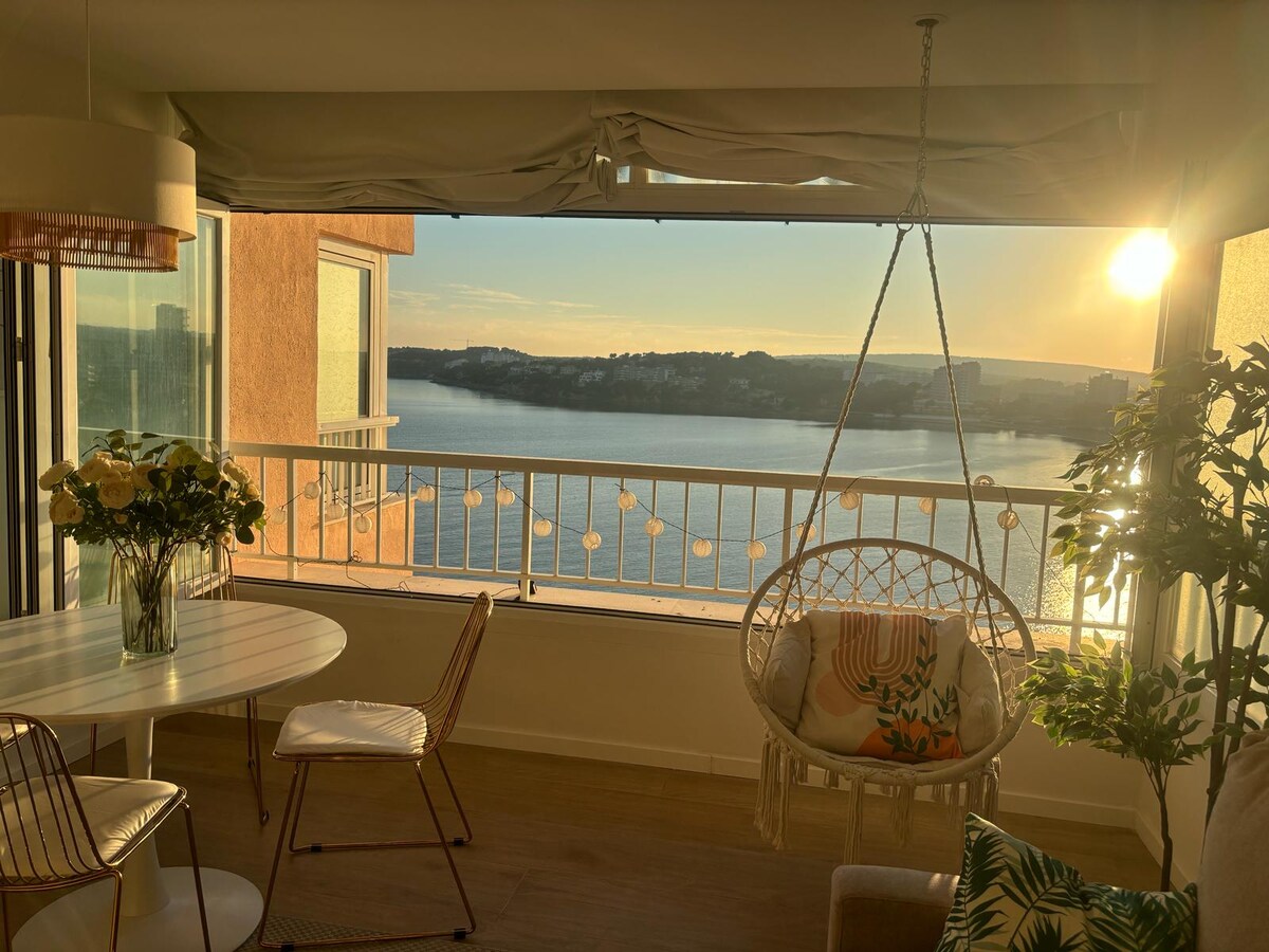 A spacious living area features a hanging chair near a large glass window, offering a view of the water and sunset. A round dining table with metal chairs sits beside a potted plant, enhancing the serene atmosphere created by natural light.