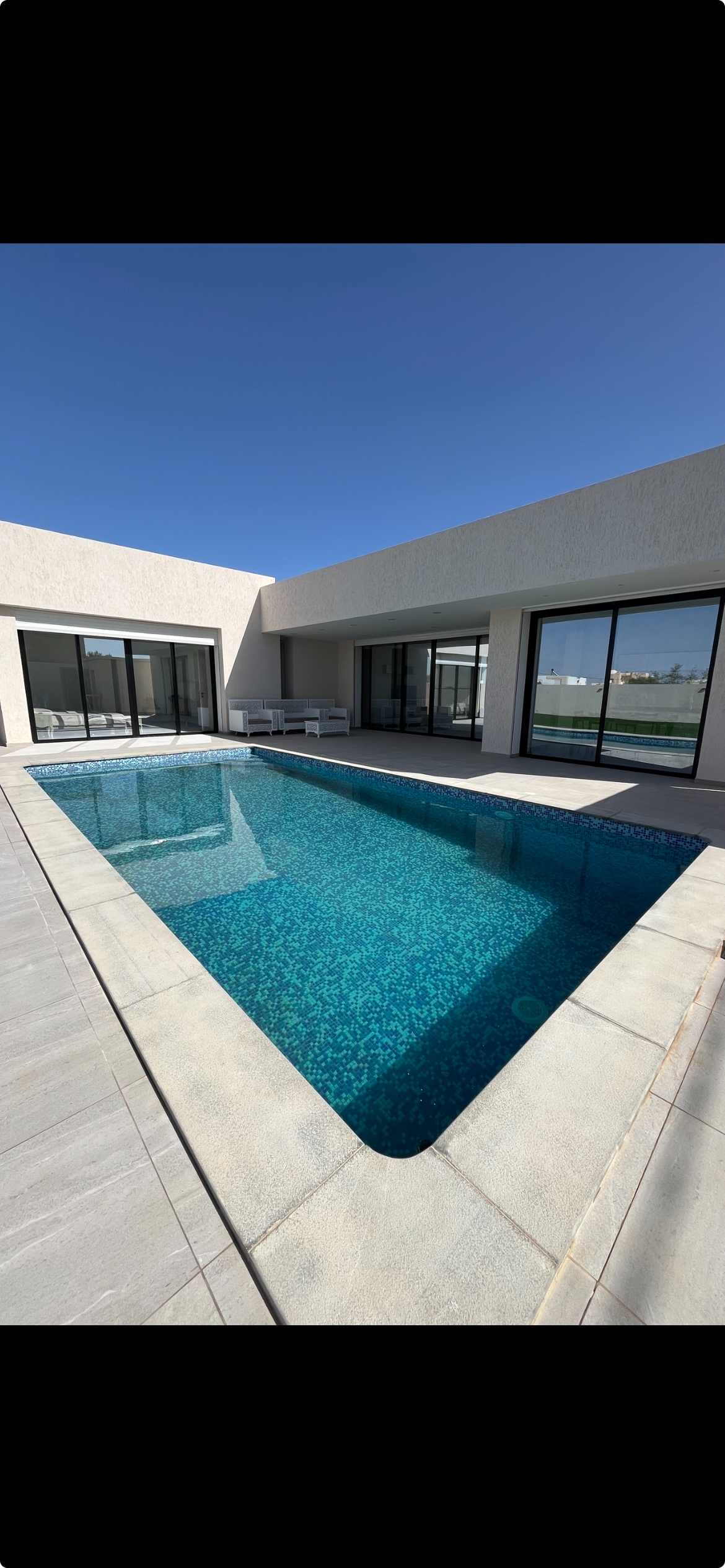 A private swimming pool is featured in the center of an outdoor area, reflecting clear blue skies above. Surrounding the pool, spacious seating areas are visible through expansive glass doors, creating a seamless connection between indoor and outdoor living.