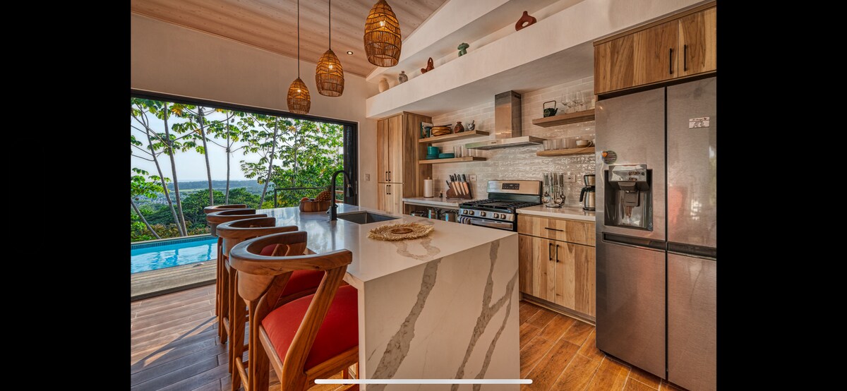 A modern kitchen features a large island with a sleek marble countertop, surrounded by wooden stools with cushions. Open shelving showcases plants and tableware, while stainless steel appliances are positioned alongside wooden cabinetry. A wall of glass offers views of the outdoor pool and surrounding greenery.