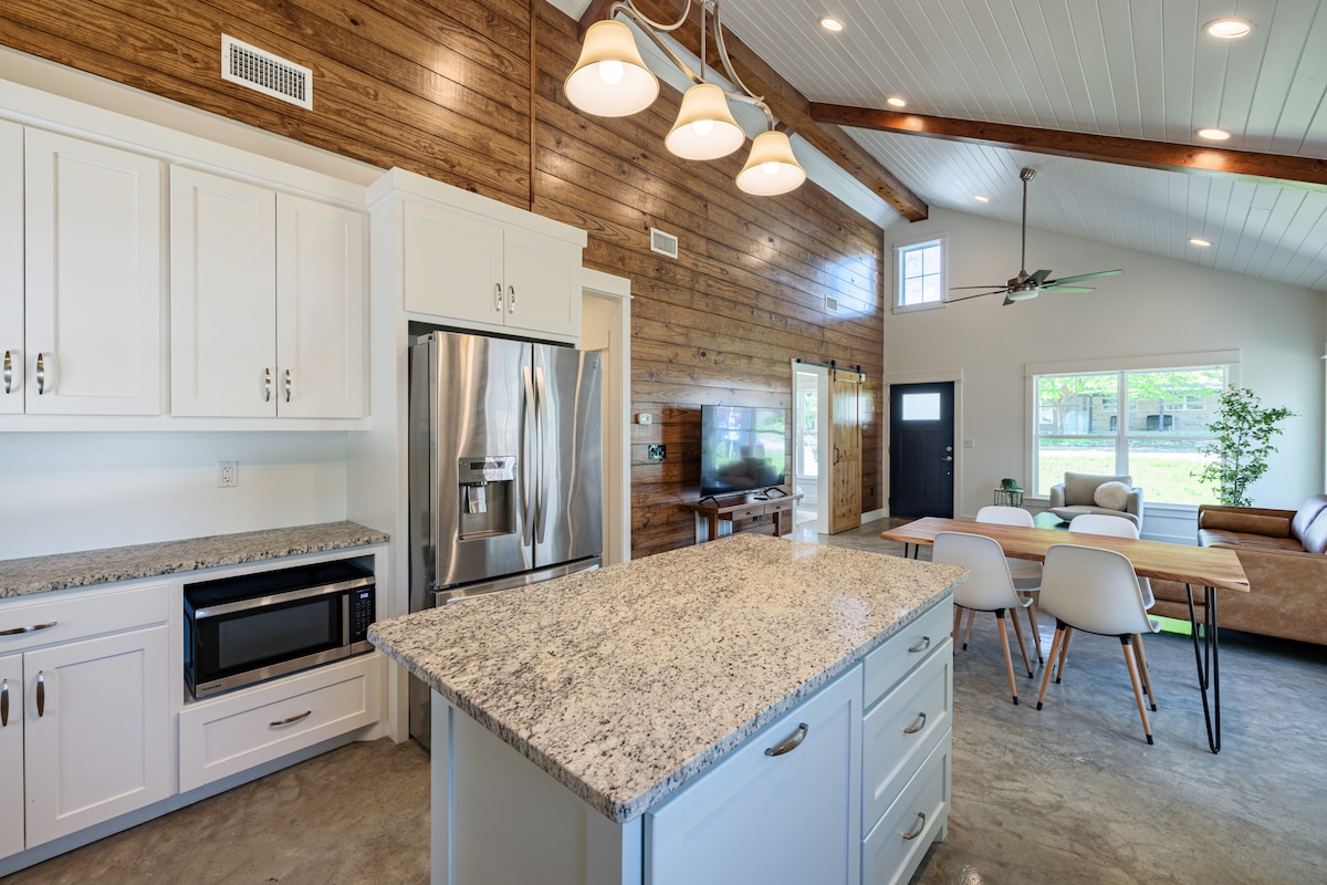 The kitchen features white cabinetry and a large island with a granite countertop. Stainless steel appliances are included, with an adjacent living area visible through a large window. A dining table with white chairs complements the open layout, enhancing functionality and natural light.