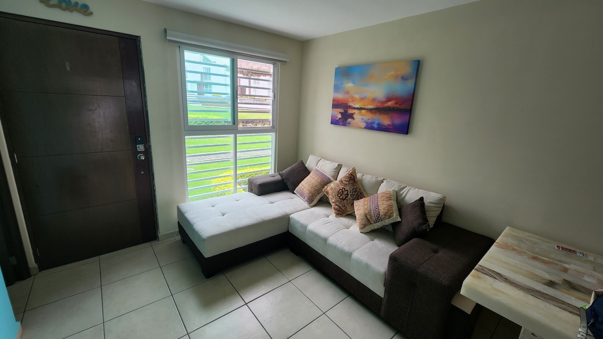 A living area features a comfortable sectional sofa adorned with decorative pillows. A small dining table is positioned nearby, while natural light filters through a large window with horizontal blinds, illuminating the neutral-toned walls and tiled flooring.