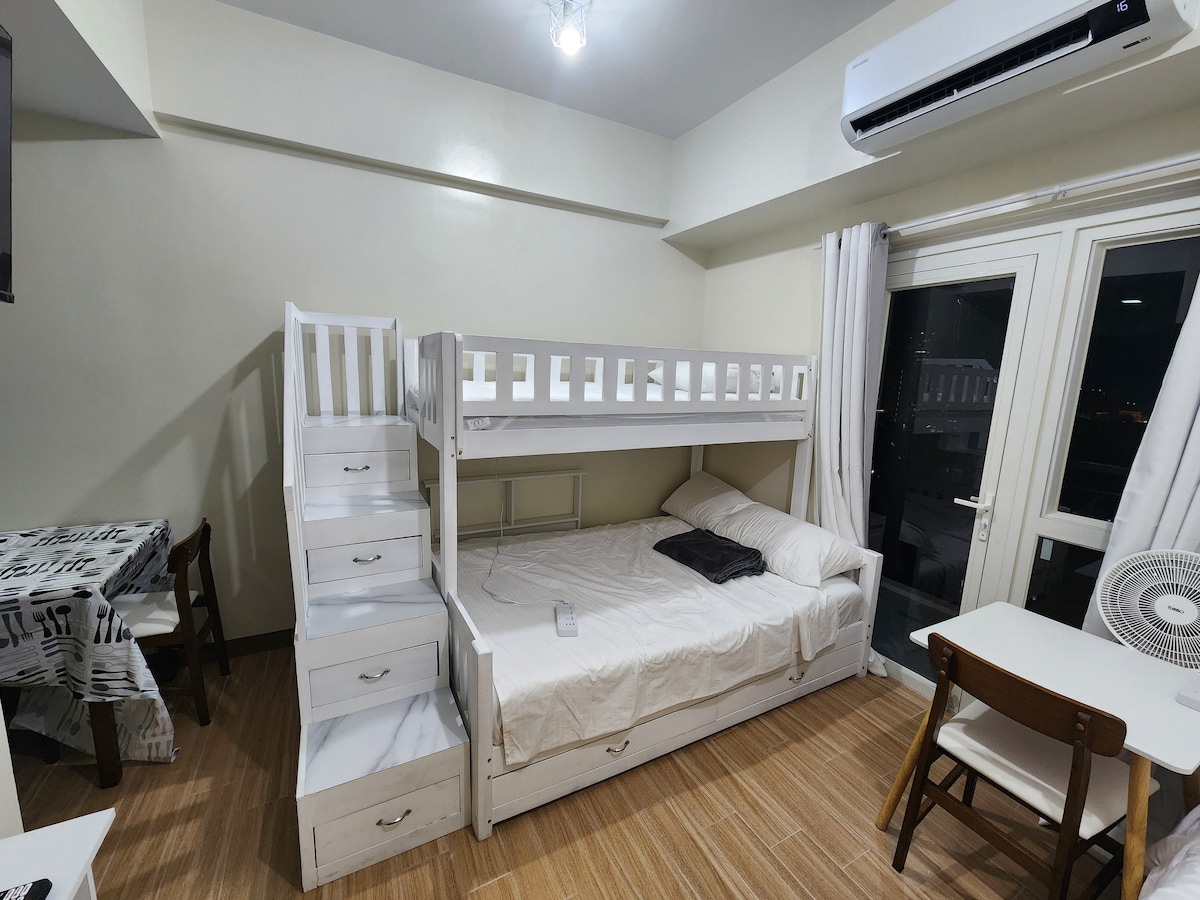 A compact room features a bunk bed with drawers underneath, providing additional storage. A small table and two chairs are positioned next to a window with sheer curtains. An air conditioning unit and a fan are also visible, ensuring comfort during the stay.