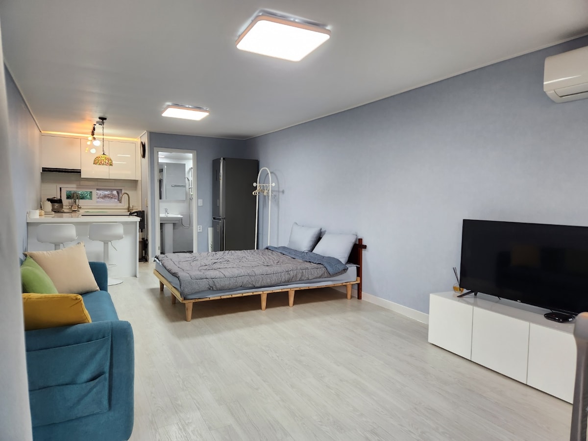 An open interior space features light-colored wood flooring and soft blue walls. A spacious bed is positioned against the wall, accompanied by two pillows. In the corner, a comfortable seating area includes a blue sofa and colorful cushions. A flat-screen television is situated on a white media console.