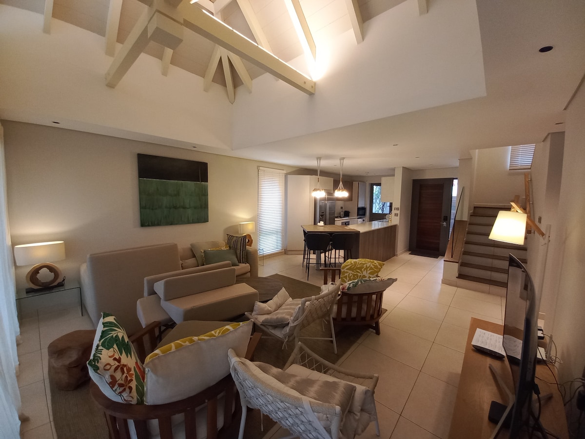 An open living area is characterized by high ceilings and natural light. Relaxing seating is arranged around a coffee table, with a modern kitchen visible in the background. Soft colors and simple decor create a calming environment, inviting family gatherings.