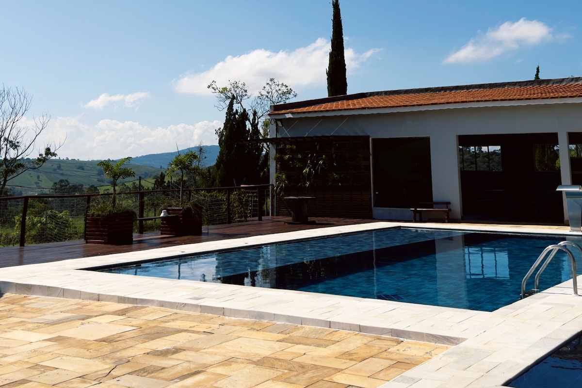 A serene outdoor area features a spacious swimming pool surrounded by a sunlit patio. The pool's clear water reflects the blue sky above, while lounge chairs are positioned nearby. The backdrop includes lush greenery and gently rolling hills, enhancing the tranquil setting.