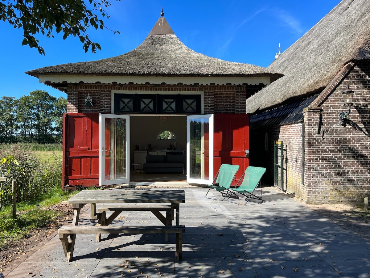 The exterior of the thatched-roof coach house is shown, featuring open double doors that lead to a welcoming interior. Two green lounge chairs are placed on the patio, alongside a wooden picnic table. Surrounding greenery adds to the tranquil setting.