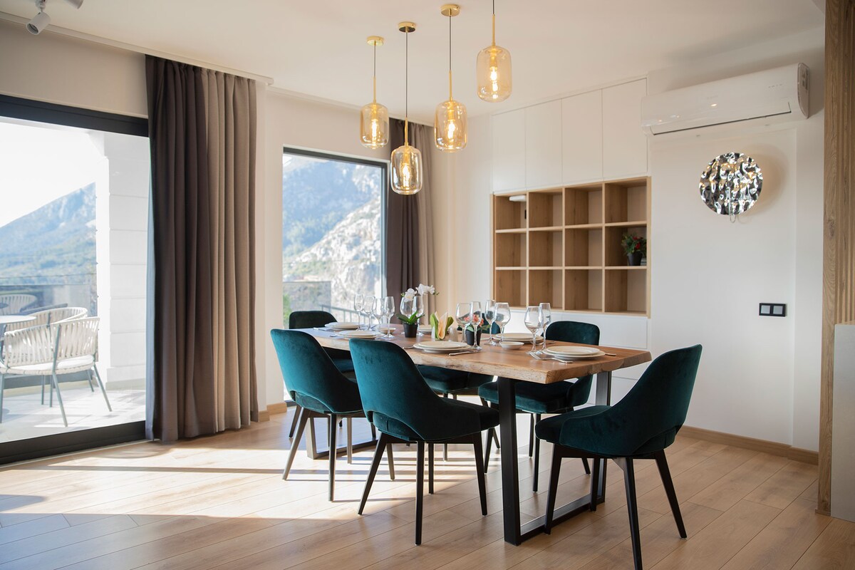 A modern dining area features a wooden table surrounded by six green upholstered chairs. Stylish pendant lights hang overhead, illuminating the space. Large glass doors provide a view of the outdoor balcony and surrounding mountains, complemented by soft drapes that enhance natural light.