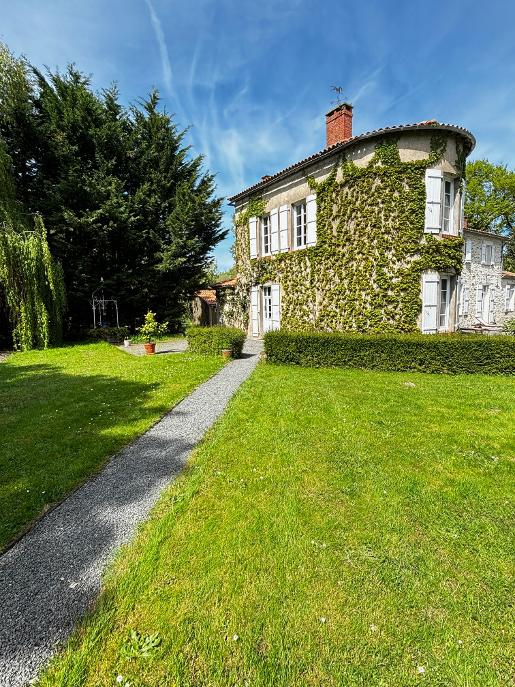 The exterior of a charming house is shown, featuring lush green vines covering a rounded turret. A well-maintained yard is visible, with a gravel pathway leading to the entrance. Several trees and shrubs provide greenery in a peaceful setting under a clear blue sky.