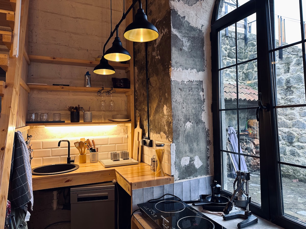 A modern kitchen area features wooden shelving and a countertop with a sink and stovetop. Industrial-style pendant lights illuminate the space. Large windows offer a view of the stone exterior, enhancing the connection to the surrounding environment.